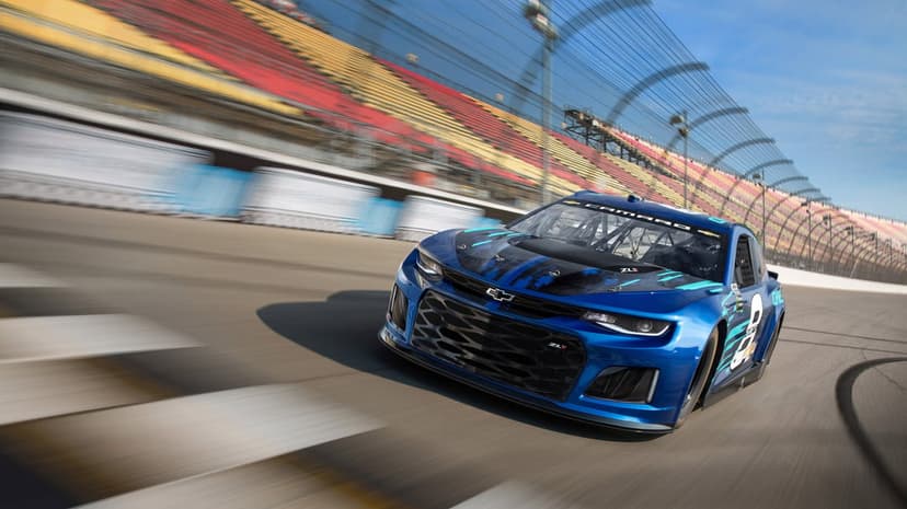 Blue Chevrolet Camaro ZL1 race car speeding on an oval track with motion-blurred grandstands and blue sky.