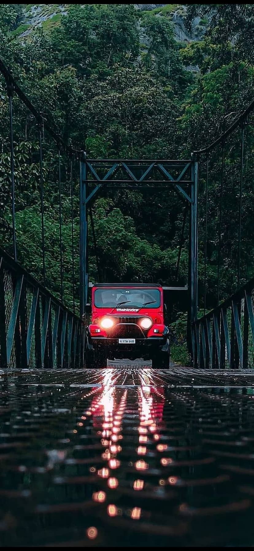 Red Mahindra Thar SUV with glowing headlights parked on a damp metallic bridge inside a dense green forest.
