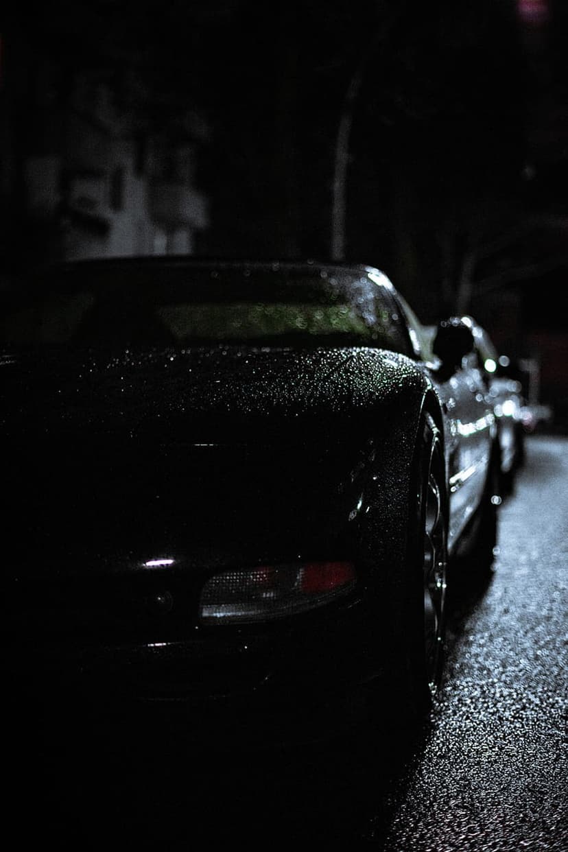 Black sportscar with raindrops on a wet asphalt street at night, featuring a glowing red taillight.