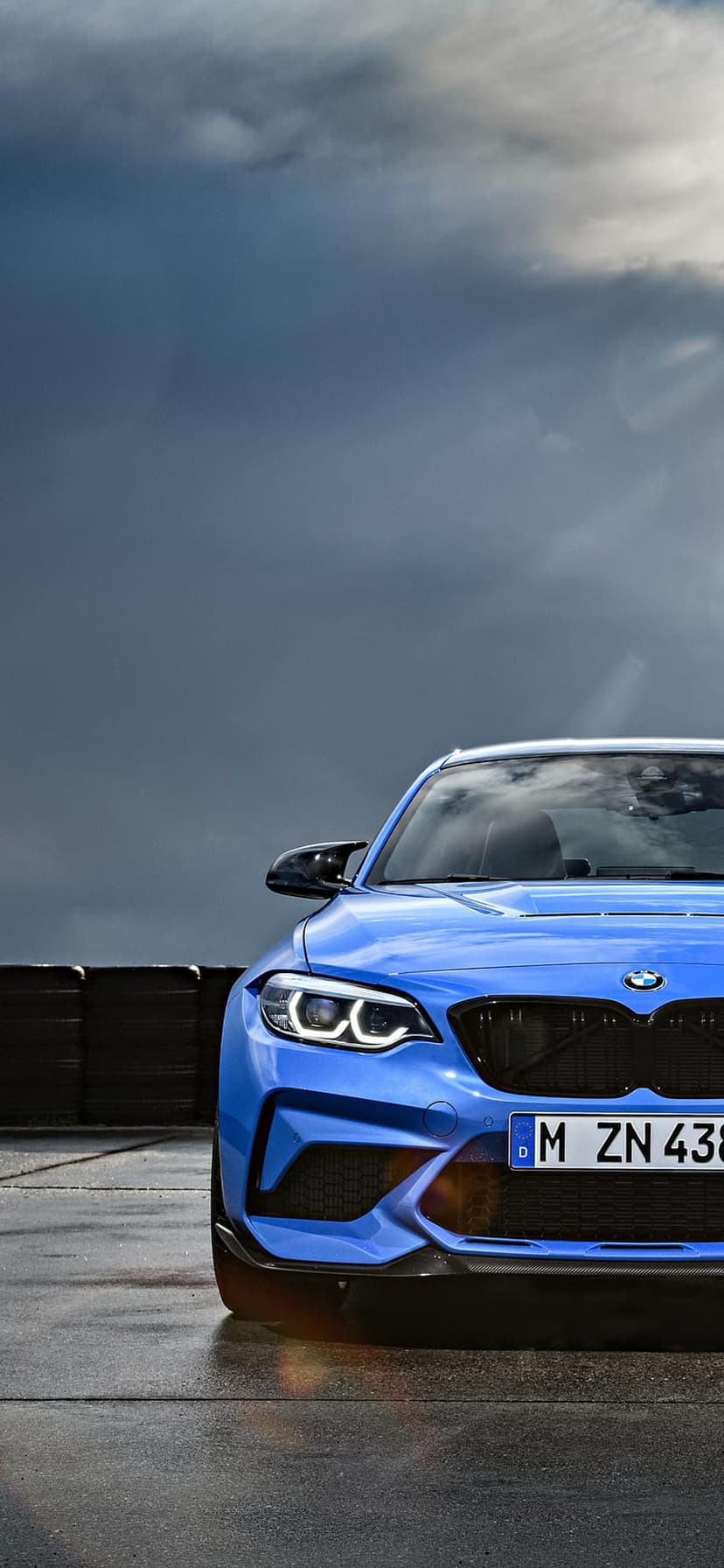 Blue BMW M2 sports car on wet asphalt with stacked tires and a dramatic cloudy sky in the background.