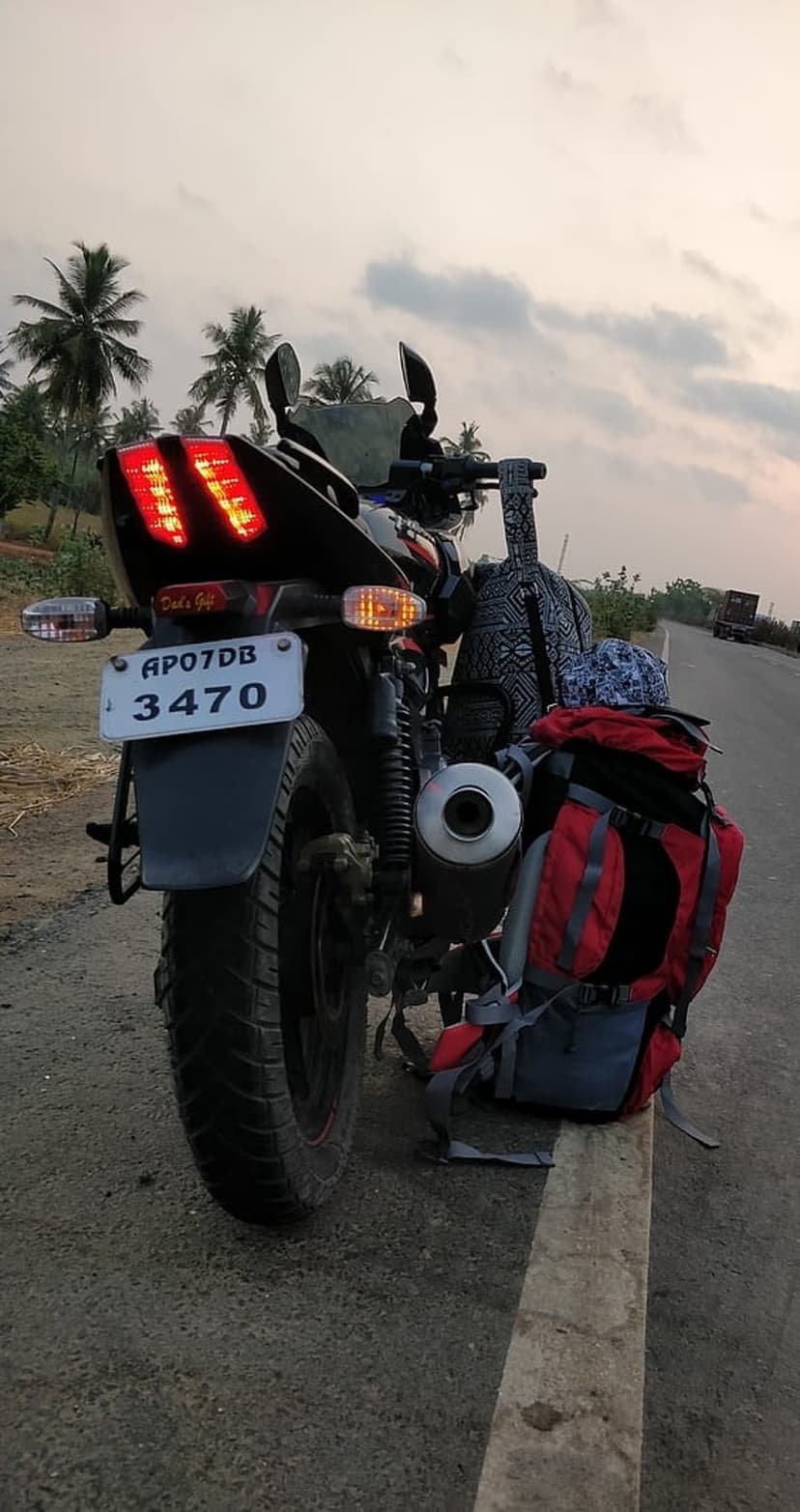 Touring motorcycle with colorful backpacks parked by palm trees under a cloudy orange sunset sky on a rural road.