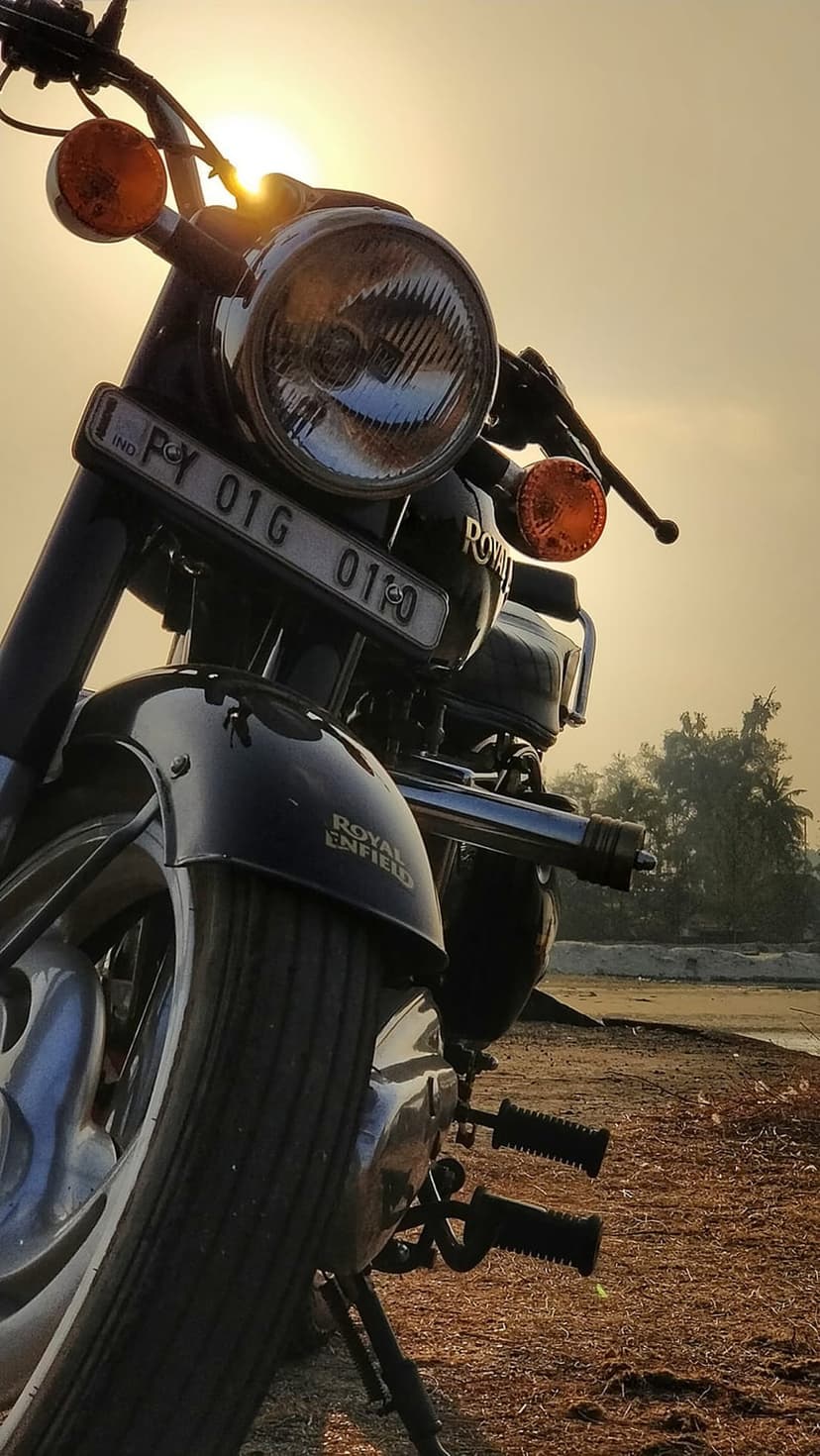 Royal Enfield motorcycle headlight and orange turn signals glowing under warm sunset backlighting in an outdoor setting.