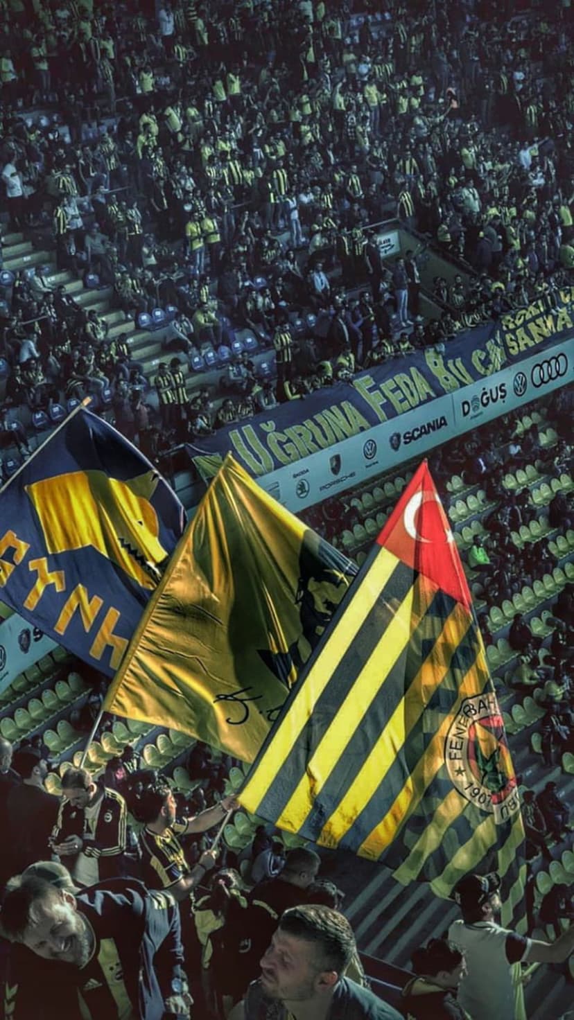 Fenerbahçe fans wave yellow and navy blue flags in a packed football stadium alongside a Turkish national flag.