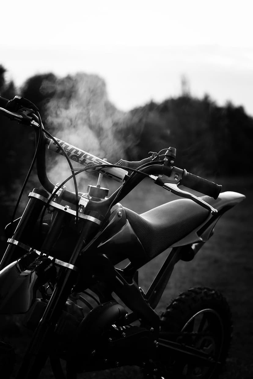 Dirt bike handlebars and a textured seat are framed by rising engine smoke in a monochrome outdoor setting.