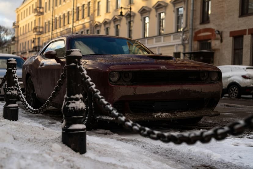Maroon Dodge Challenger parked on a snowy city sidewalk behind thick steel chains during a winter sunset.