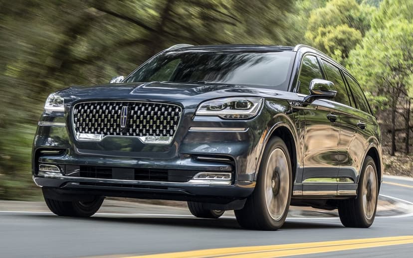 Dark charcoal Lincoln Aviator SUV driving on a curved road through a dense green forest with motion blur.