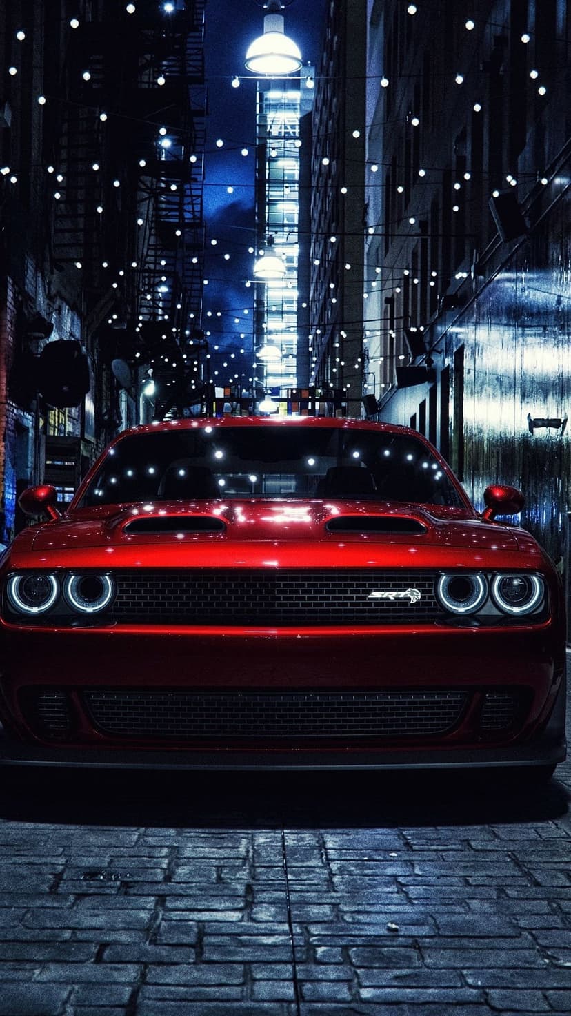 Red Dodge Challenger parked in a narrow alleyway under hanging string lights on a textured cobblestone street.
