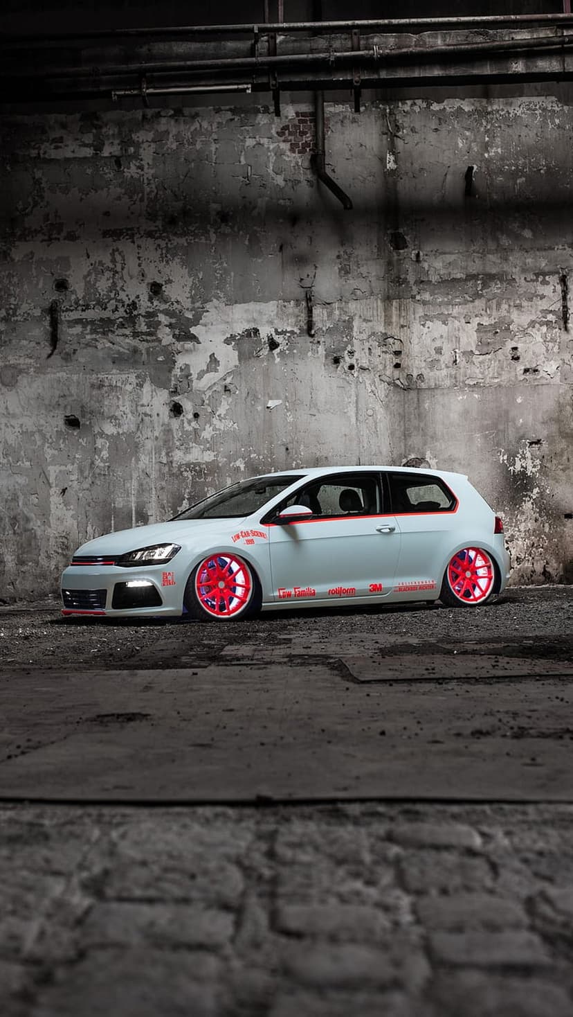White Volkswagen Golf GTI with red wheels and lowered suspension parked against a textured grey concrete wall.