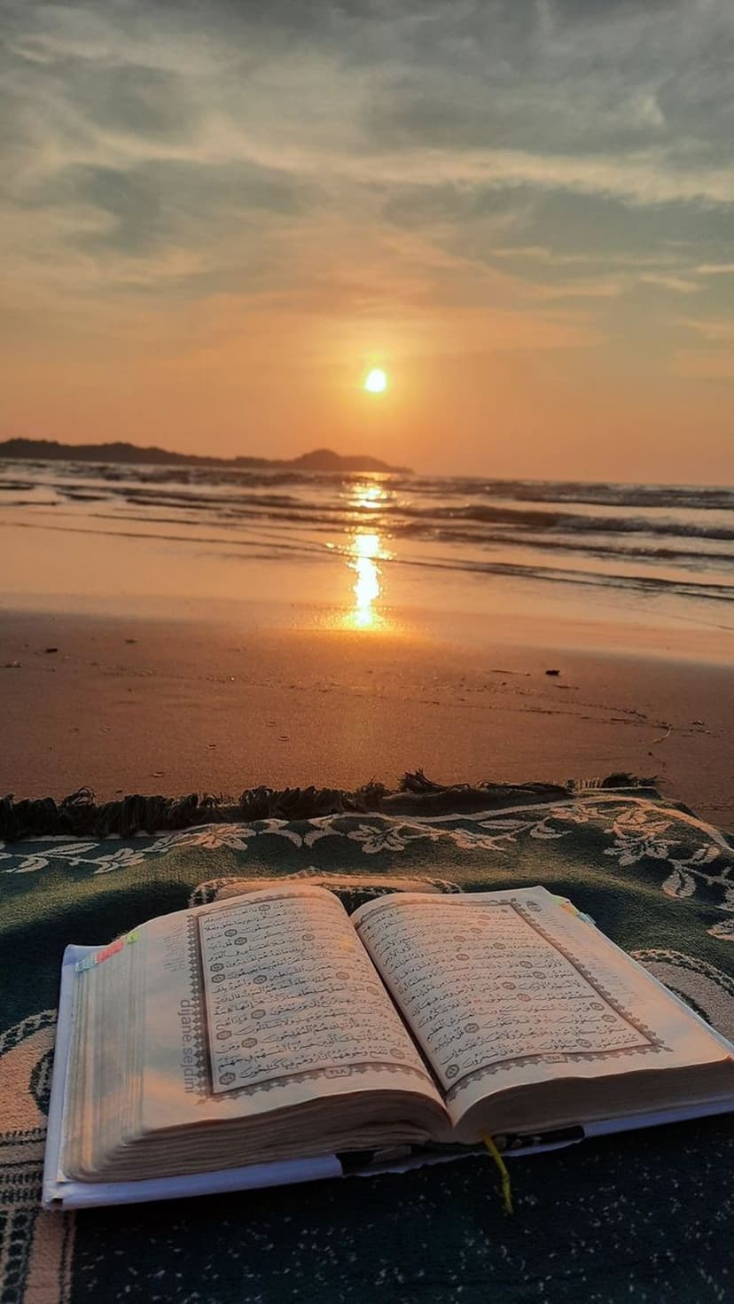 Quran on a prayer rug sits on sandy beach at sunset while gentle ocean waves approach the sacred book.