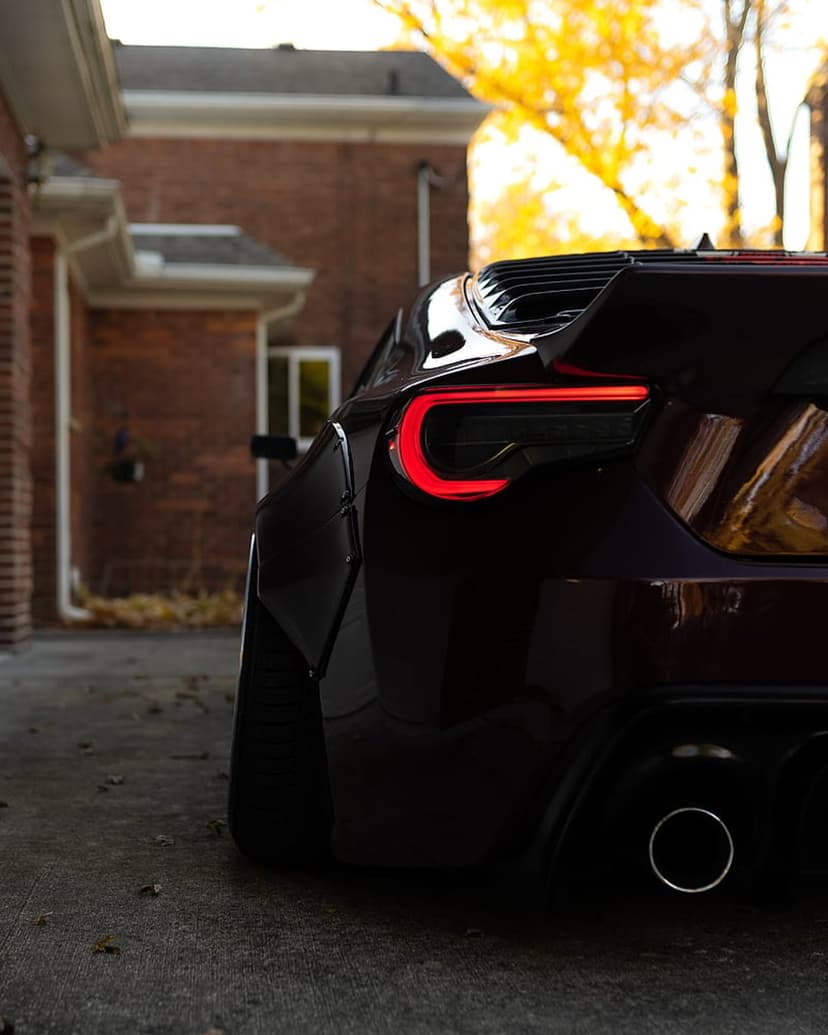 Dark red sports car rear with glowing C-shaped LED taillights, a large spoiler, and autumn leaves behind it.