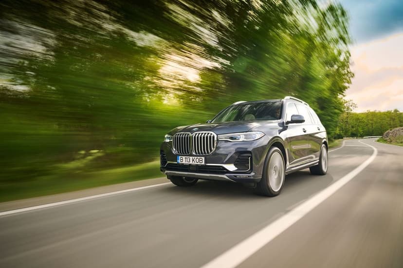 Dark grey BMW X7 luxury SUV speeding on a winding forest road with motion blur and lush green trees.