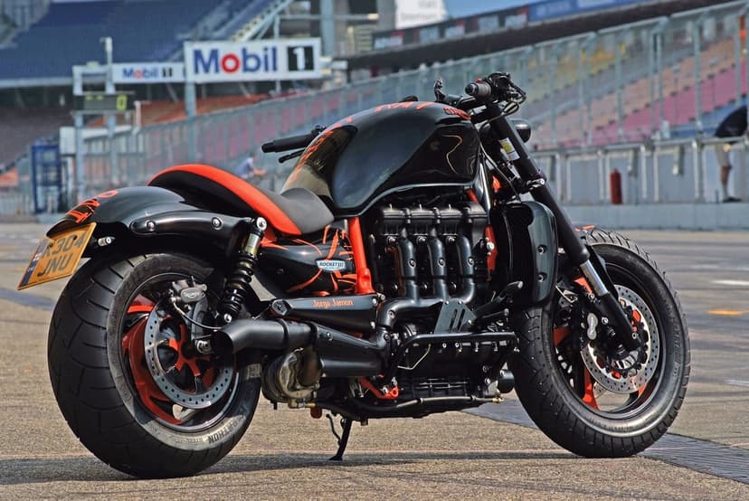 Custom black and orange Triumph Rocket III motorcycle parked on a racing circuit with a blurred stadium background.