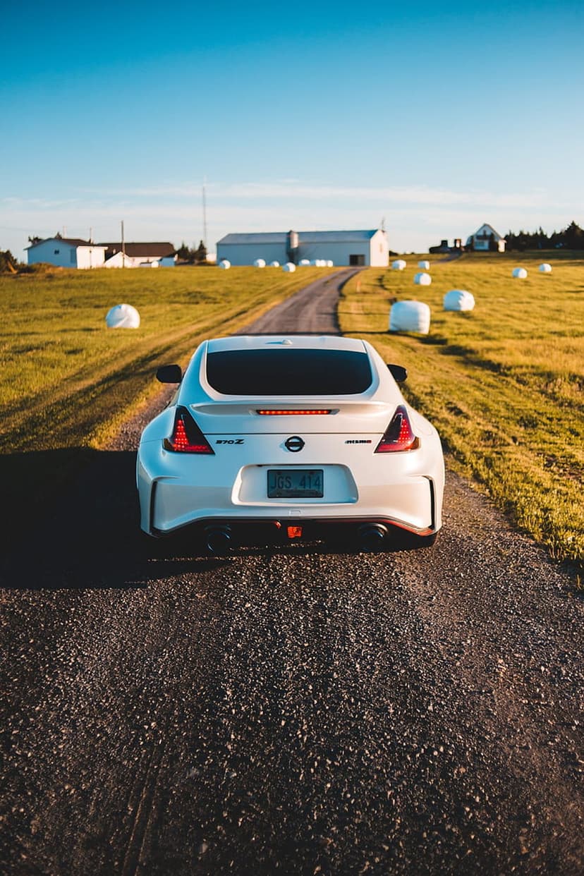 White Nissan 370Z rear view parked on a gravel path near large hay bales and green fields under a clear blue sky.
