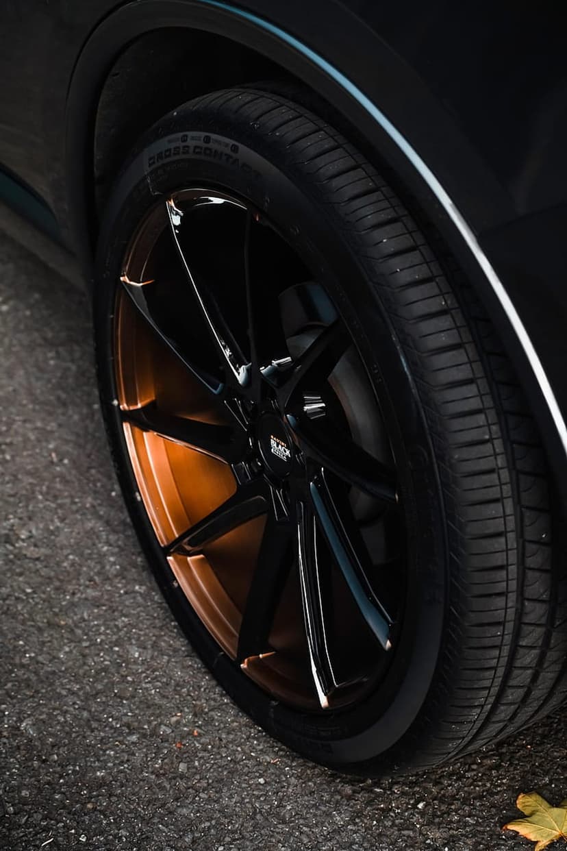 Black multi-spoke car wheel with bronze inner accents rests on dark asphalt with visible tire tread detail.