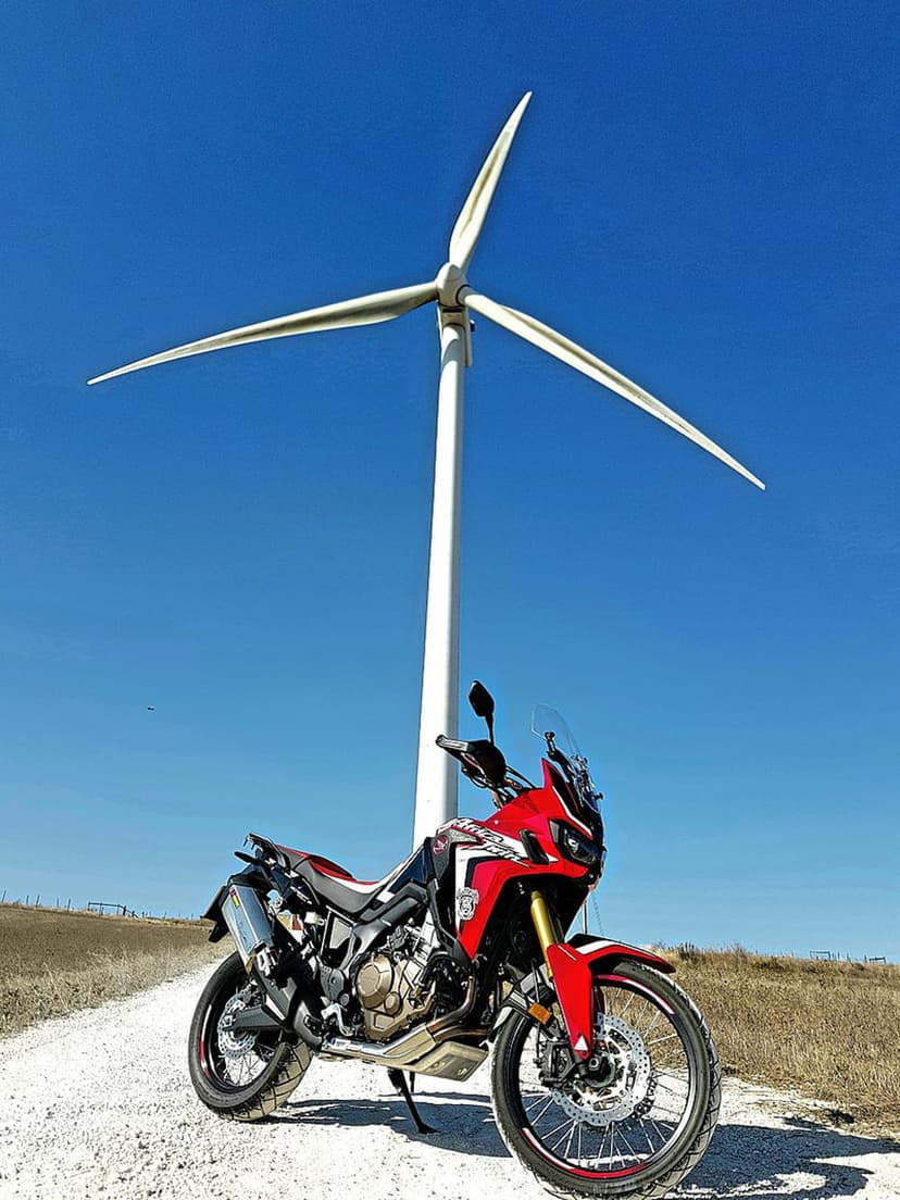 Red adventure motorcycle parked on a gravel road beneath a white wind turbine against a bright blue sky.