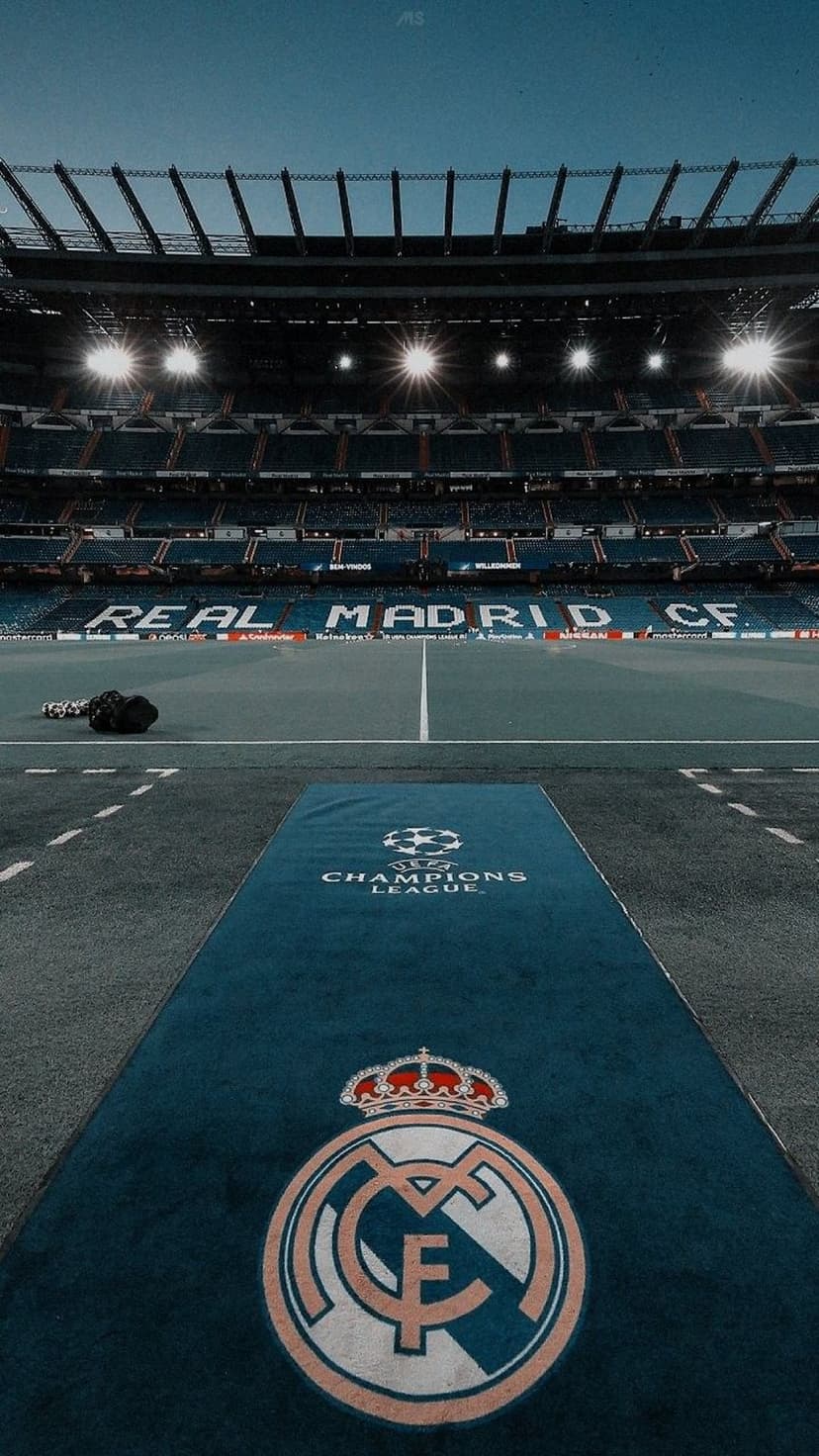 Real Madrid crest and Champions League logo on a blue carpet leading to an illuminated stadium at night.