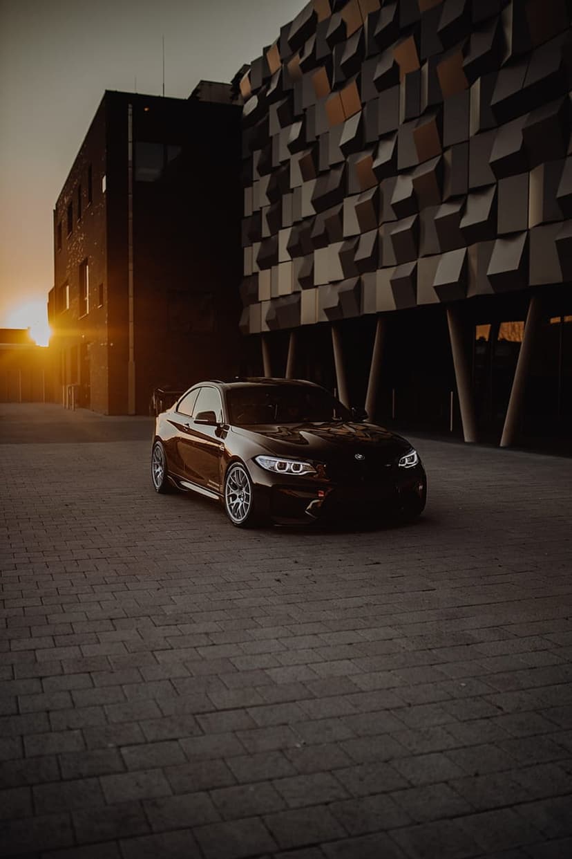 BMW M2 coupe parked in front of a textured modern building with golden sunset light and long shadows on the ground.