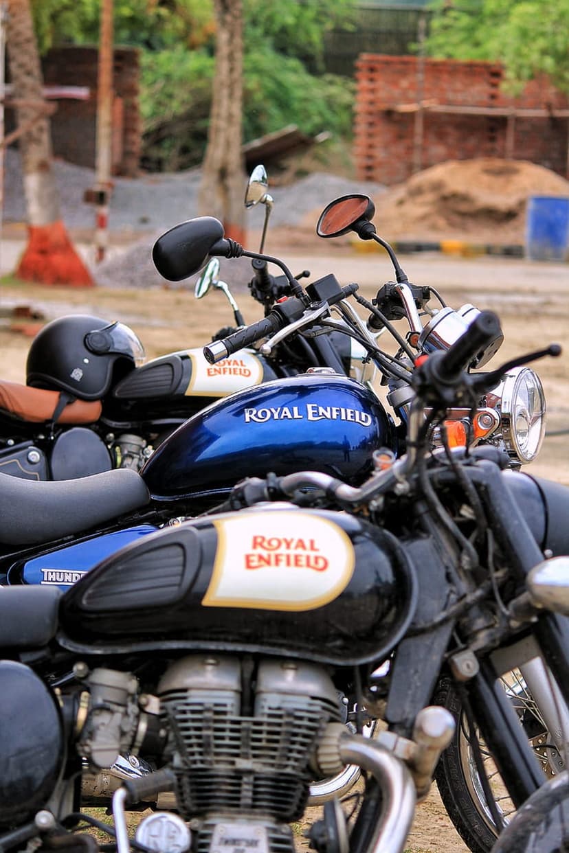 Blue and black Royal Enfield fuel tanks parked on gravel with blurred brick walls and trees in the background.