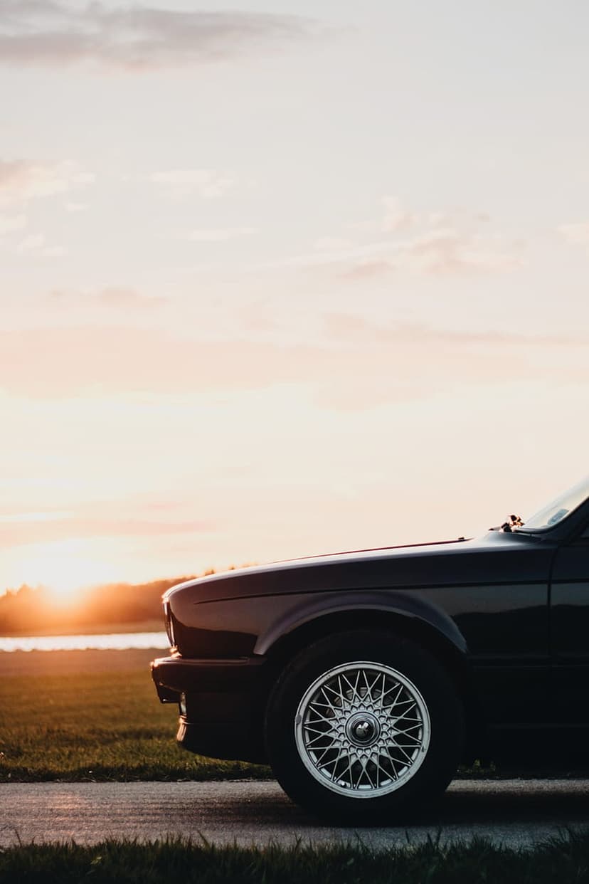 BBS mesh wheel on a black BMW E30 at sunset with golden light hitting the car fender and a grassy field behind.