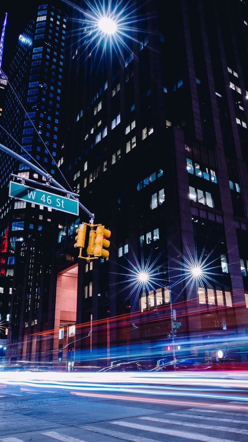 Blue and red light trails zoom past a West 46 St sign and skyscrapers in a night New York City street scene.