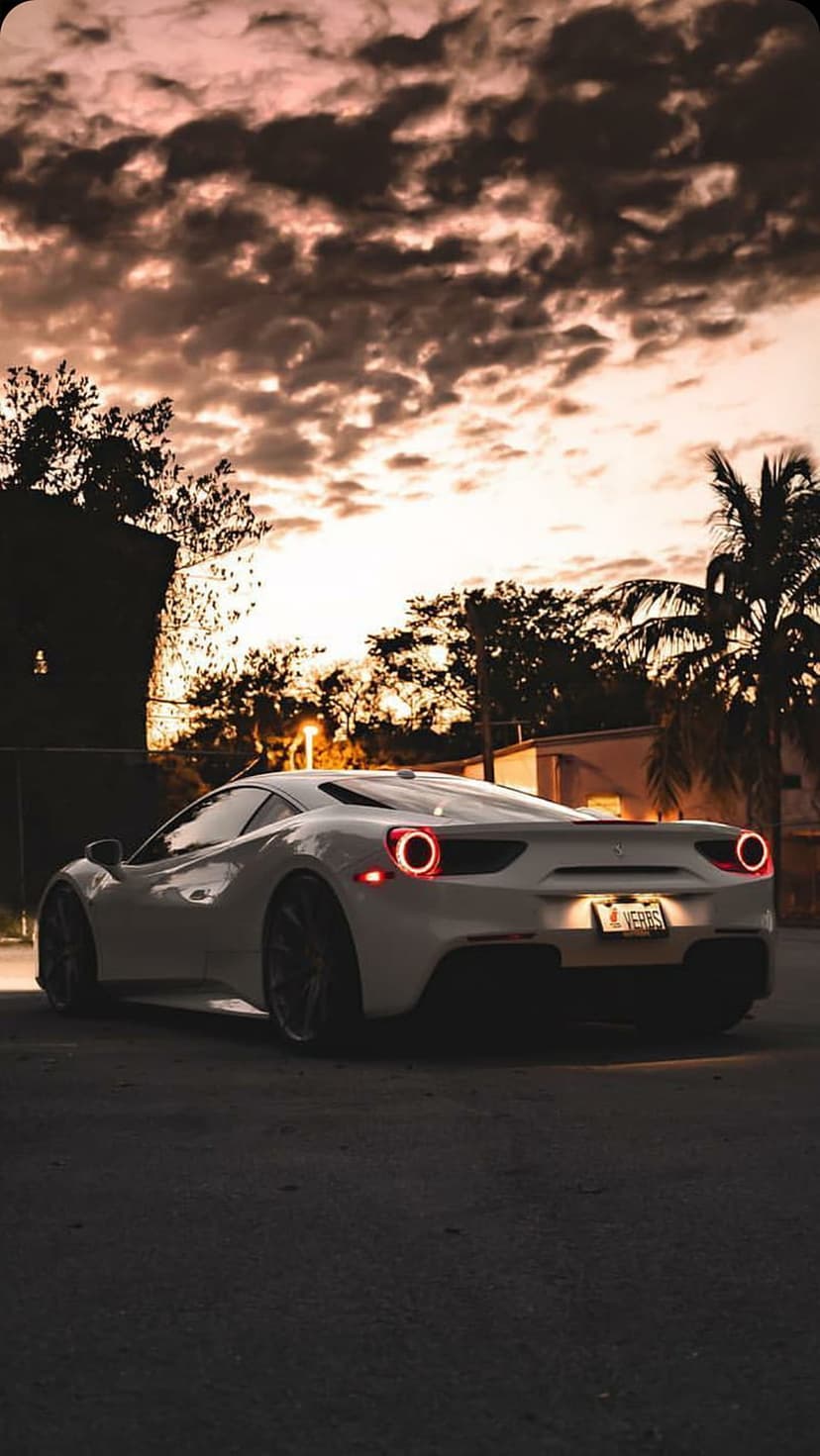 White Ferrari supercar rear view parked on asphalt against a dramatic orange and pink sunset sky with palms.
