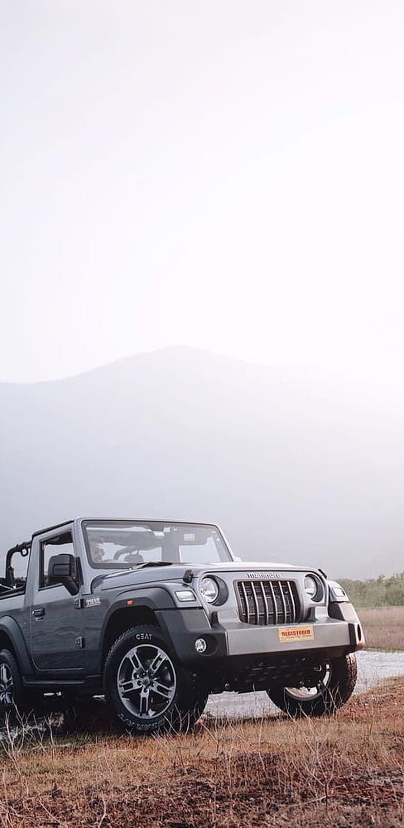 Mahindra Thar convertible SUV parked on dry grass with soft misty mountain peaks visible in the background.