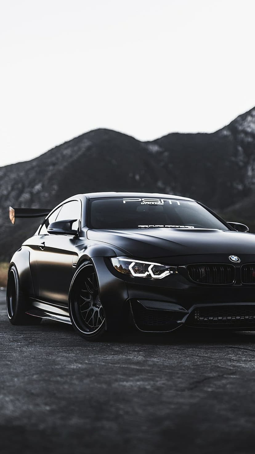 Matte black BMW M4 widebody coupe with a large rear spoiler and halo headlights on a blurred mountain road.