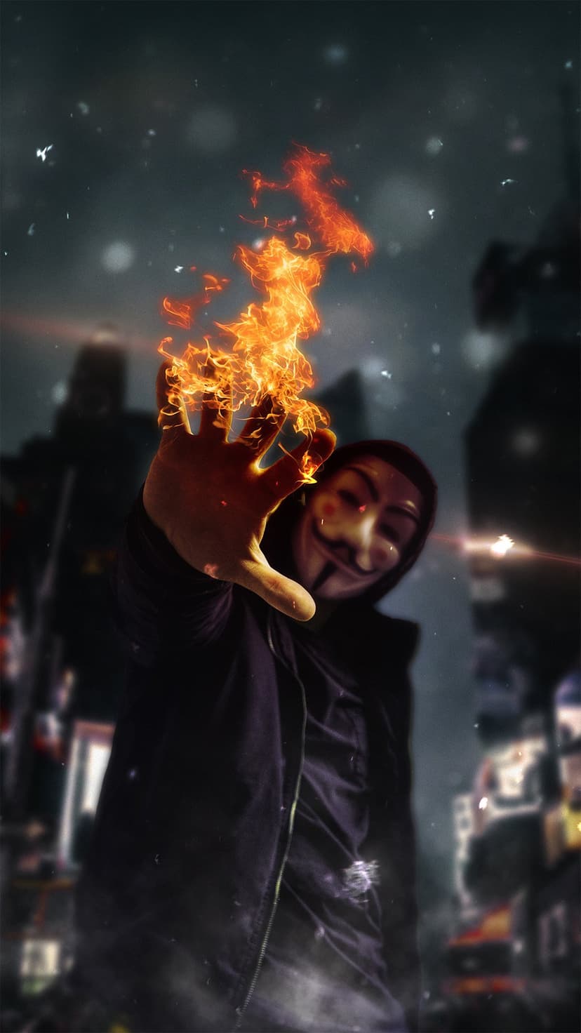 Guy Fawkes masked person holding out a palm emitting bright orange flames against a blurred dark city background.