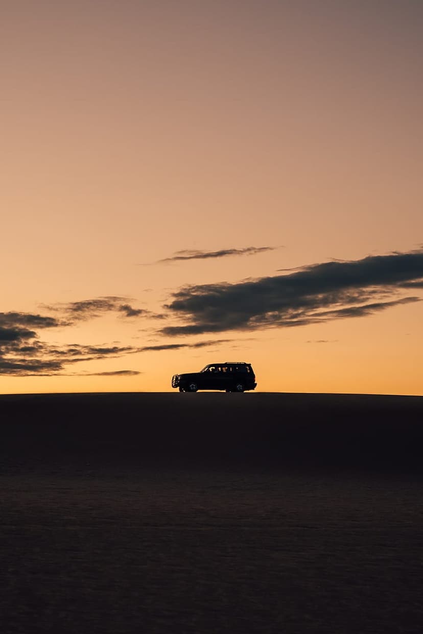 Jeep silhouette on a dark sand dune against a vibrant orange and pink sunset sky with wispy clouds.