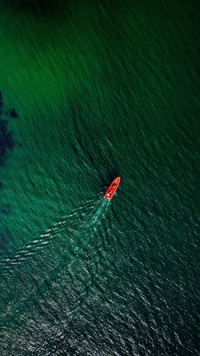 Red boat leaving a white wake on shimmering emerald and turquoise ocean water seen from a top-down view.