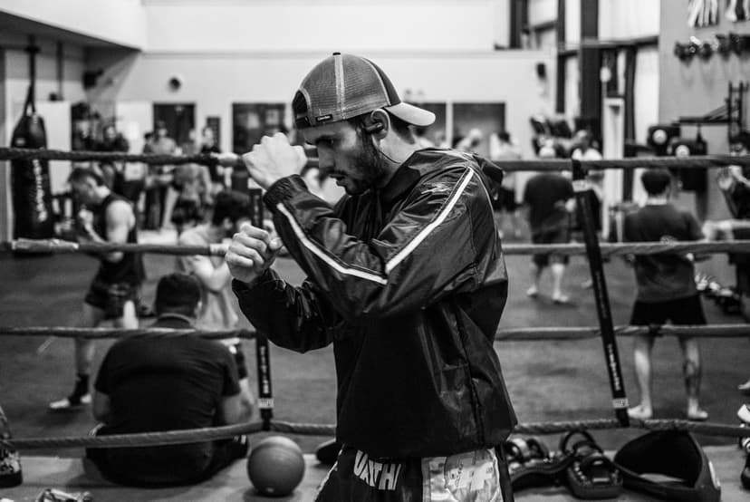 Boxer in a baseball cap maintains a defensive stance in a monochrome ring during a gym training session.
