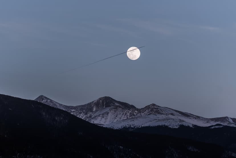 Full moon bisected by a white jet contrail over sharp, snow-dusted mountain ridges against a dark night sky.
