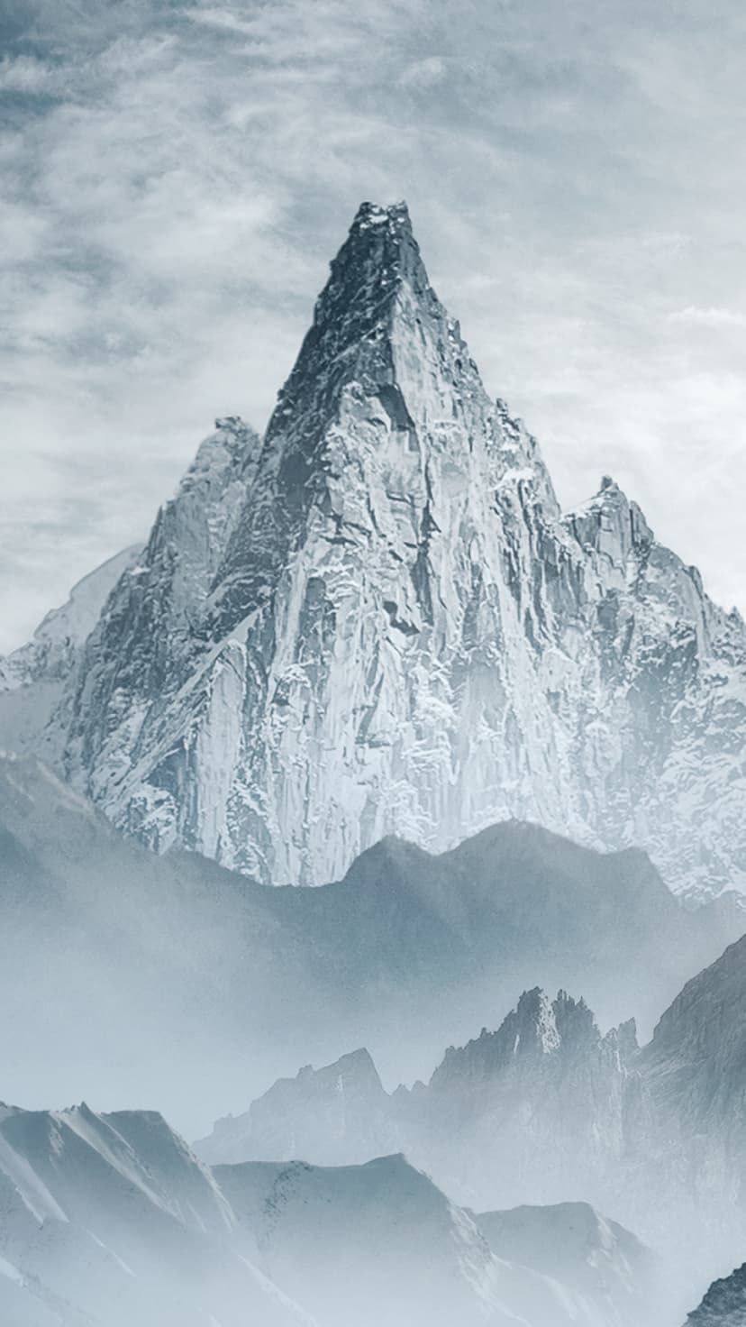 Jagged mountain peak covered in thick snow and ice rises above a layer of low-hanging mist under grey skies.