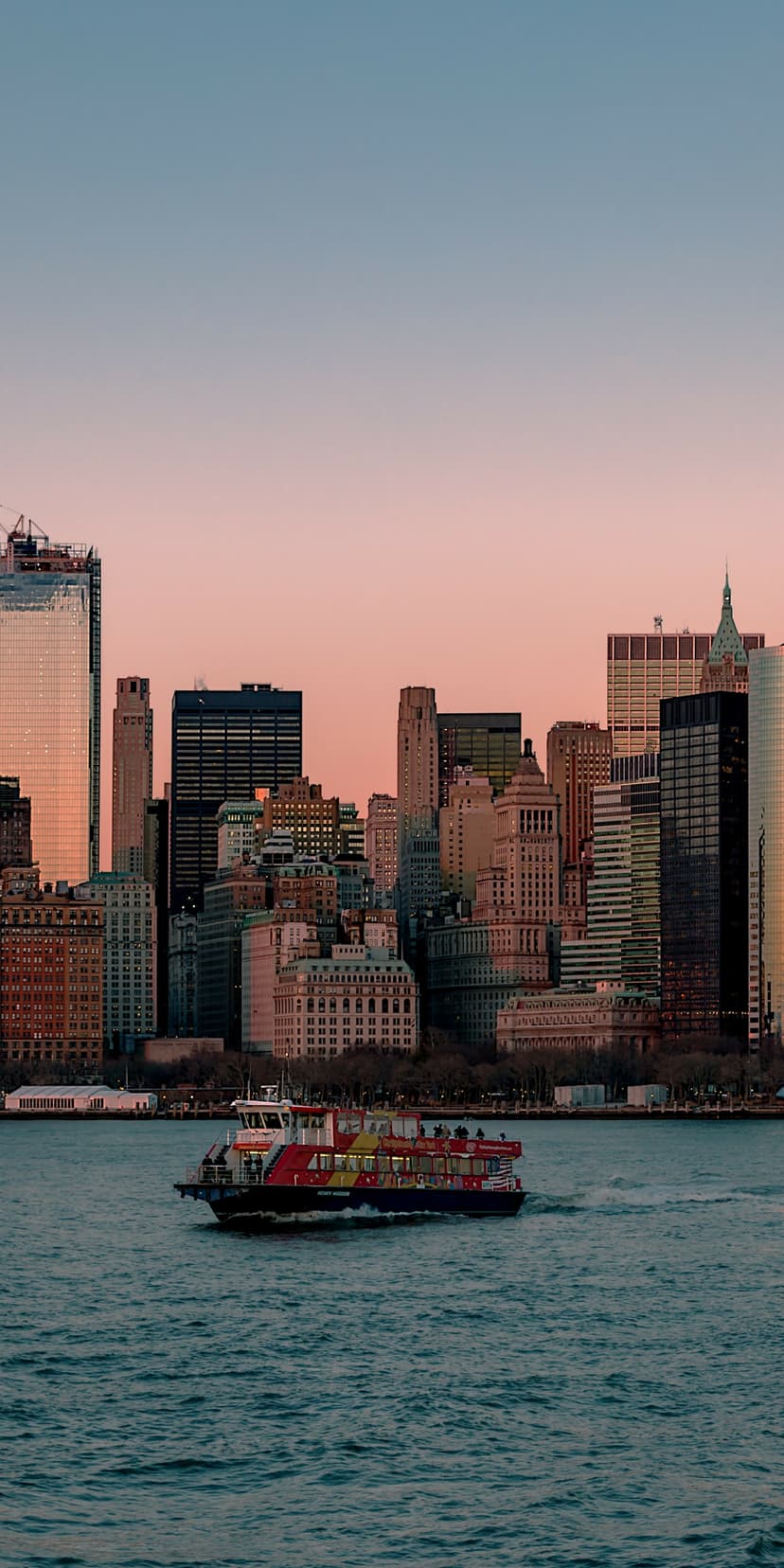 Red and yellow ferry on calm water with the Manhattan skyline and a pink sunset sky in the background.