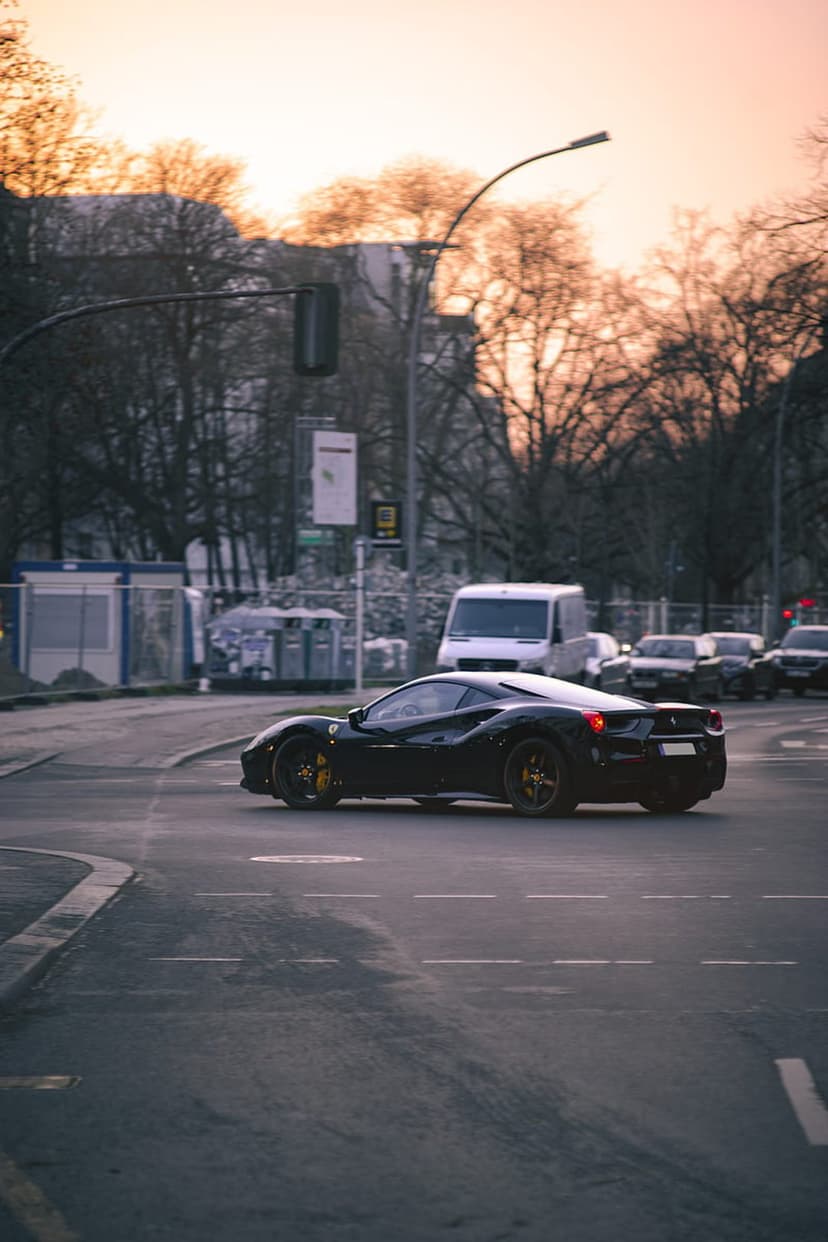 Black Ferrari sports car turns onto a city street during a vibrant orange sunset with trees in the background.