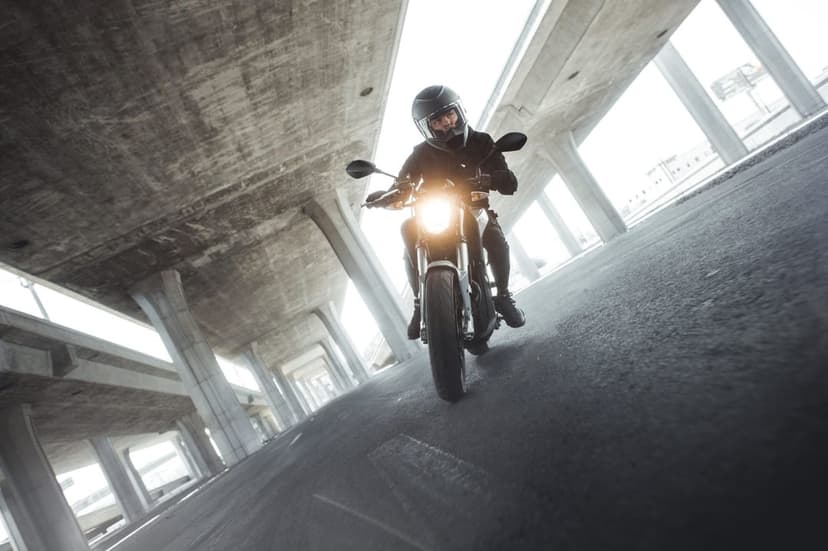 Motorcyclist on a white bike riding under a massive concrete bridge with bright headlights in an urban setting.