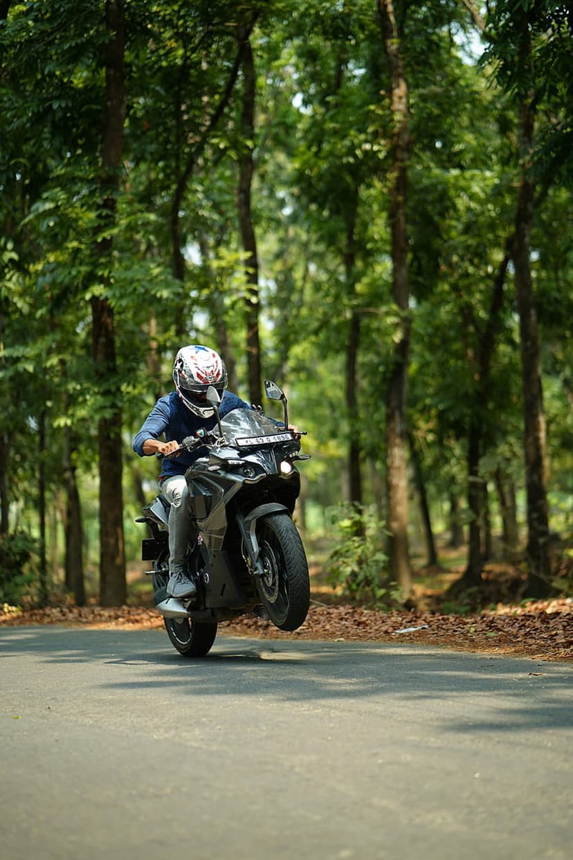 Motorcyclist in a blue shirt performs a wheelie on a black bike along a tree-lined paved road in the forest.