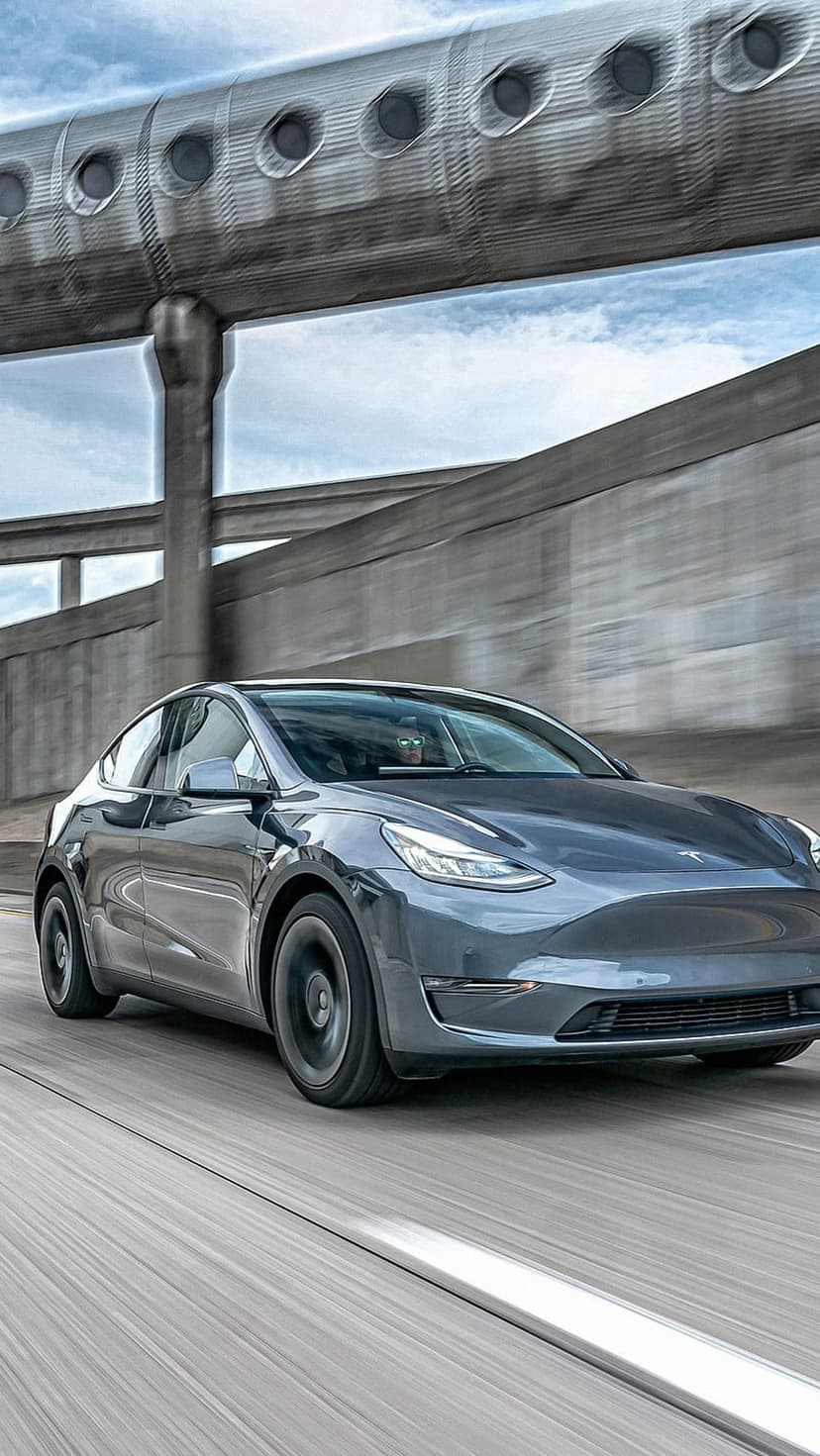 Tesla Model Y in metallic grey speeds along a highway with motion blur against a cloudy sky and concrete overpasses.