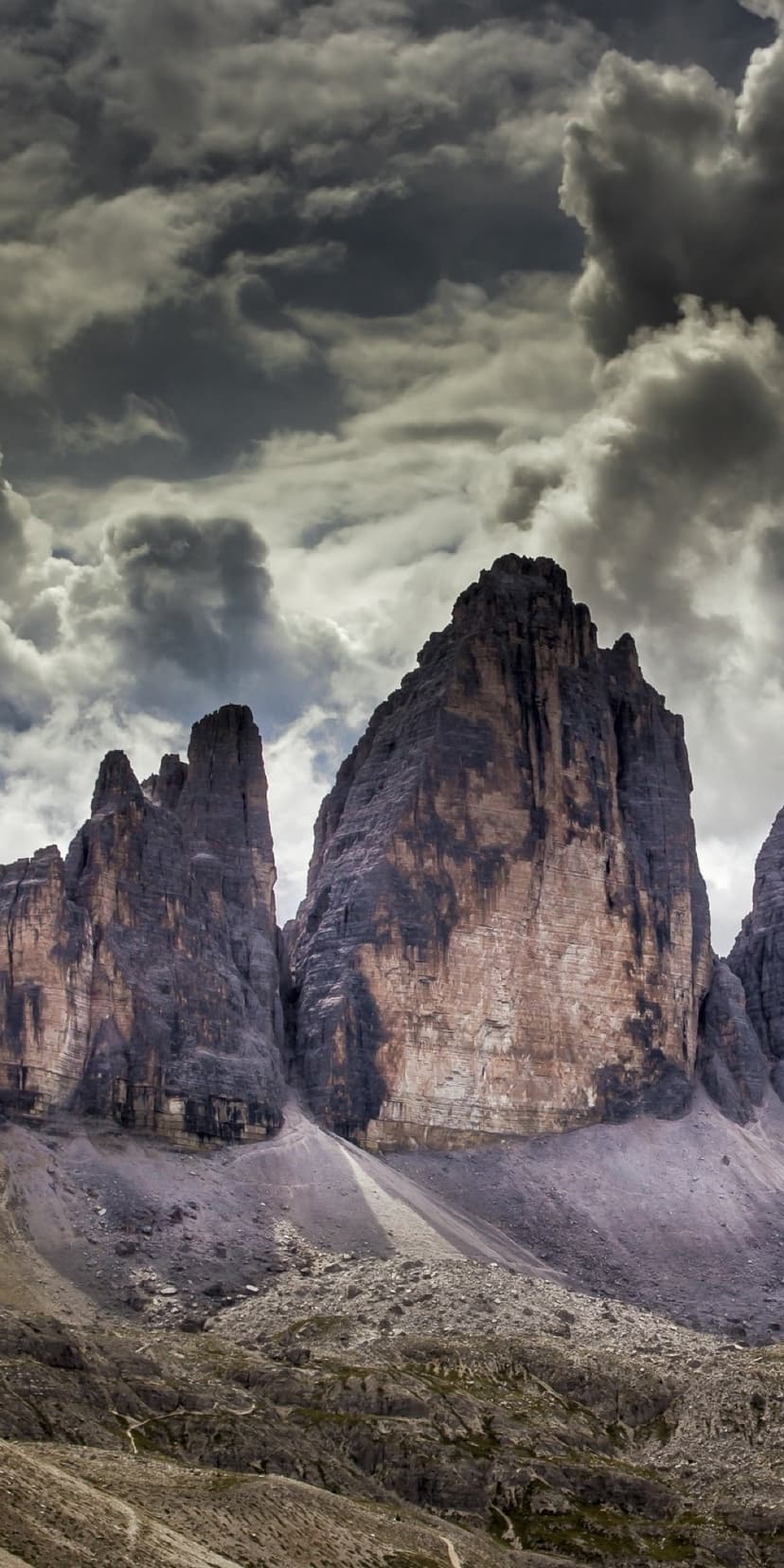 Three jagged mountain peaks rise toward a dark overcast sky in this vertical Italian Dolomite landscape.