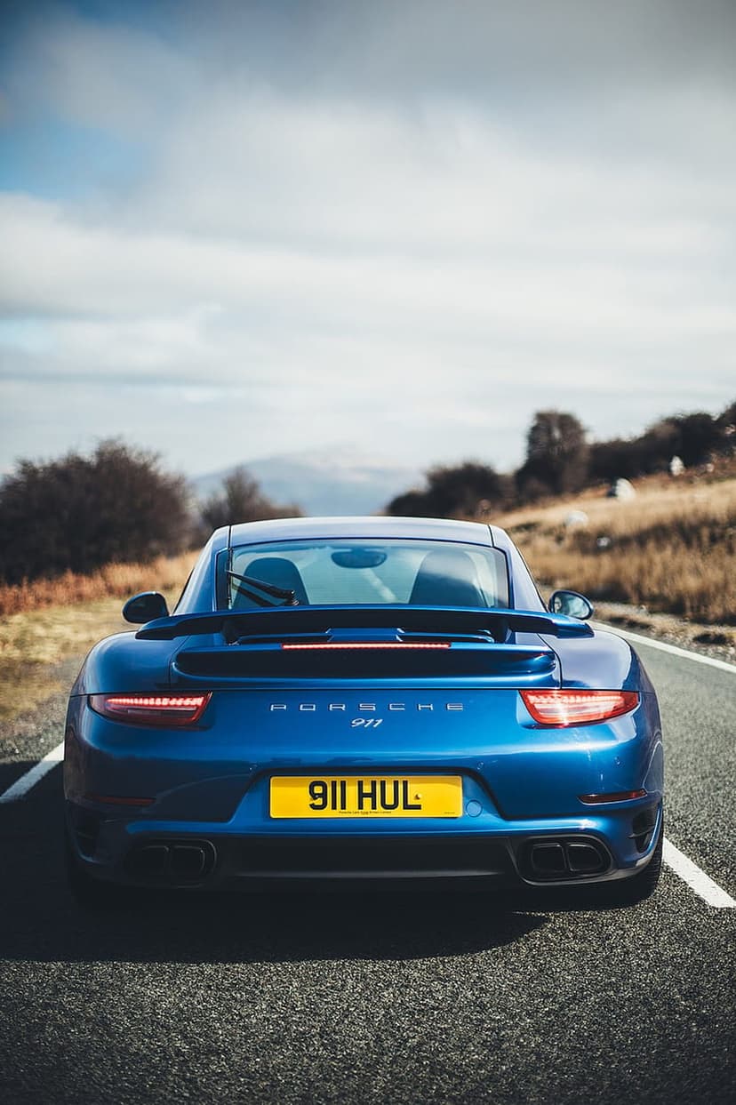 Blue Porsche 911 rear view with quad exhausts and yellow license plate on a winding asphalt road through hills.