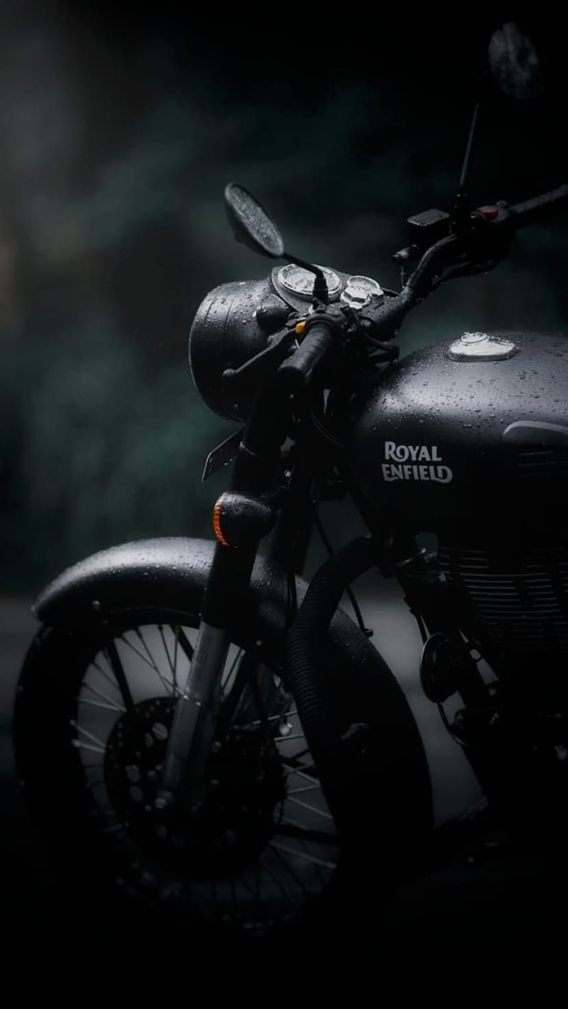 Black Royal Enfield motorcycle with water droplets on the headlight parked against a dark blurred forest.