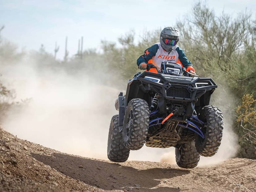 Black Polaris ATV jumping over a dirt mound in a desert with dust clouds and a rider in Klim gear.