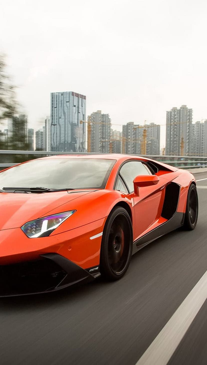 Orange Lamborghini Aventador supercar driving on an urban highway with motion-blurred skyscrapers in the background.