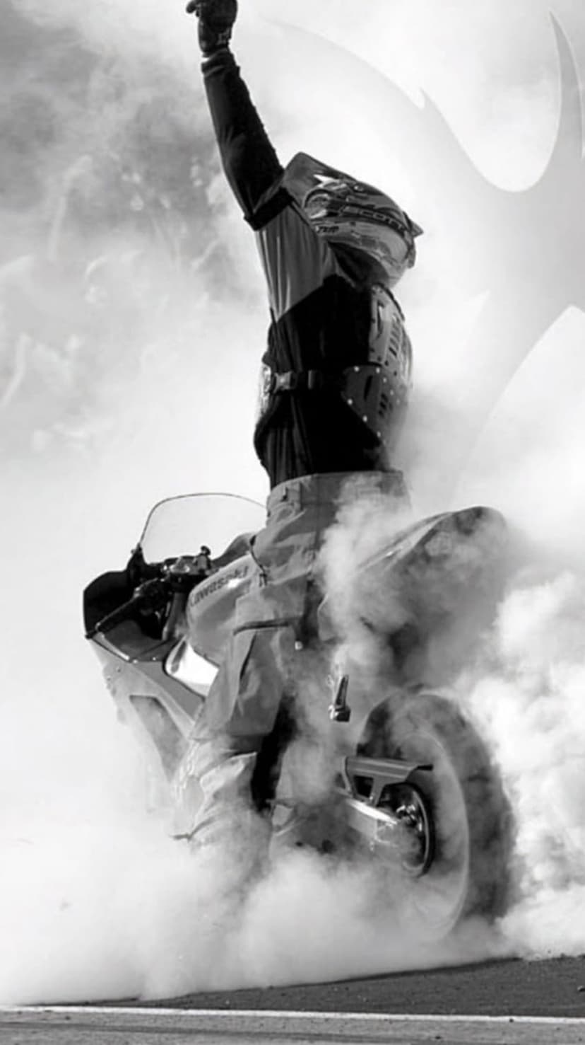 Motorcyclist in full gear performs a front-wheel lift as thick smoke billows from the ground in monochrome.