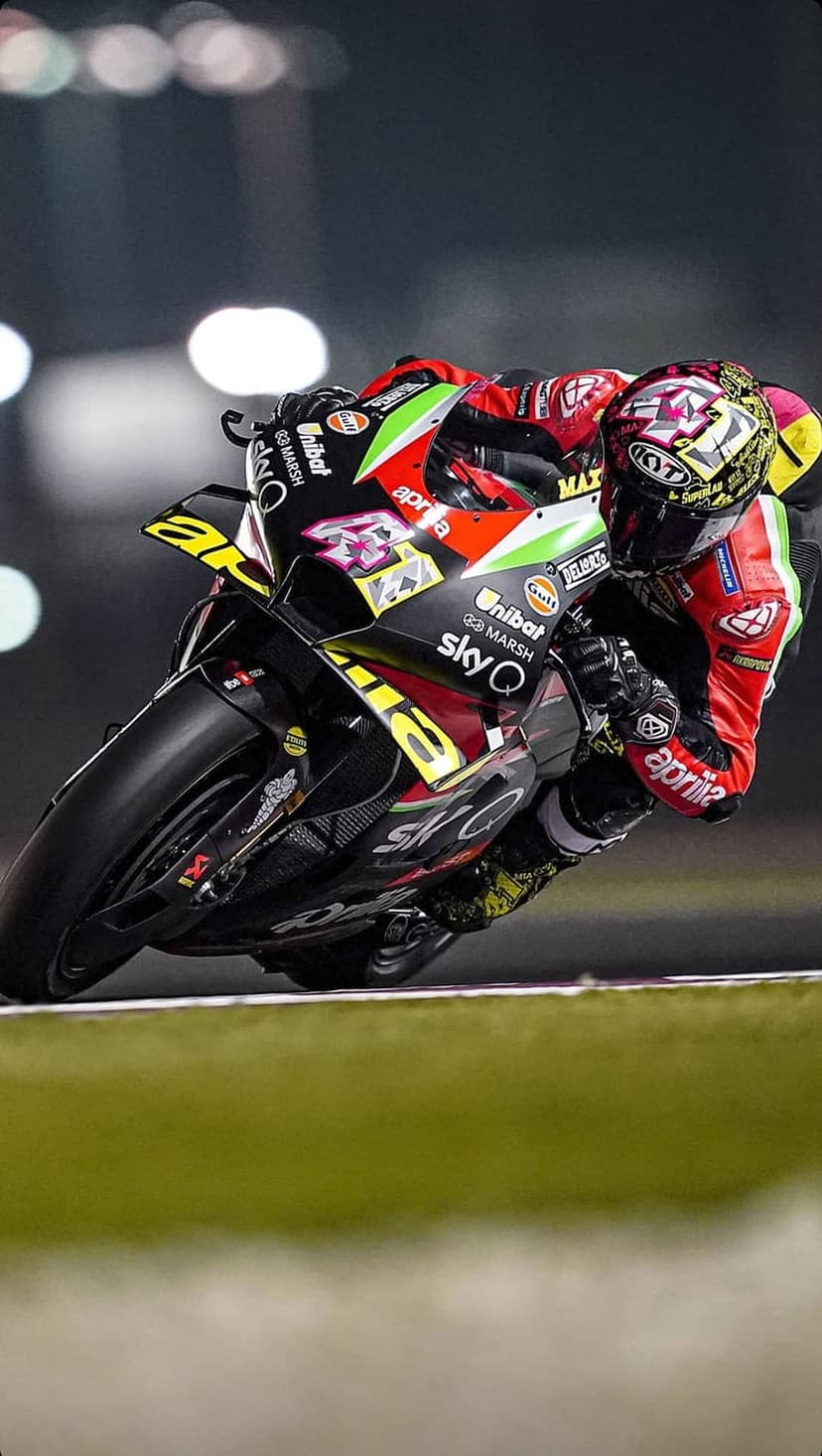 Motorcyclist in red and black racing leathers leans into a dark track curve under bright stadium lights.
