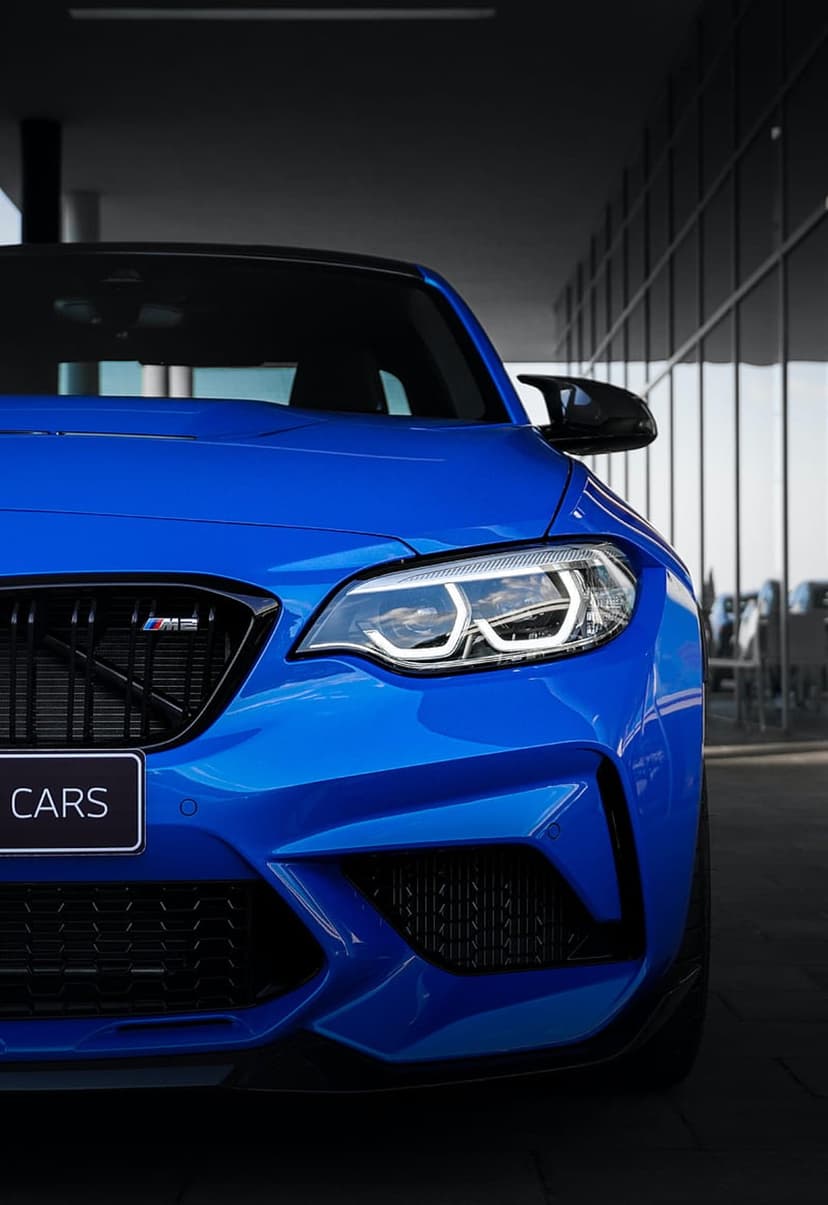 Blue BMW M2 sports car front view featuring a kidney grille and LED headlights against a glass building background.