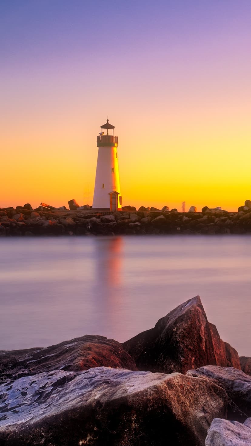 White lighthouse on dark rocks under a purple and orange sunset sky reflecting in calm coastal water.