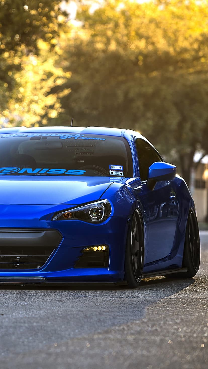 Blue custom sports car with yellow fog lights parked on asphalt under dappled sunlight in a low-angle shot.