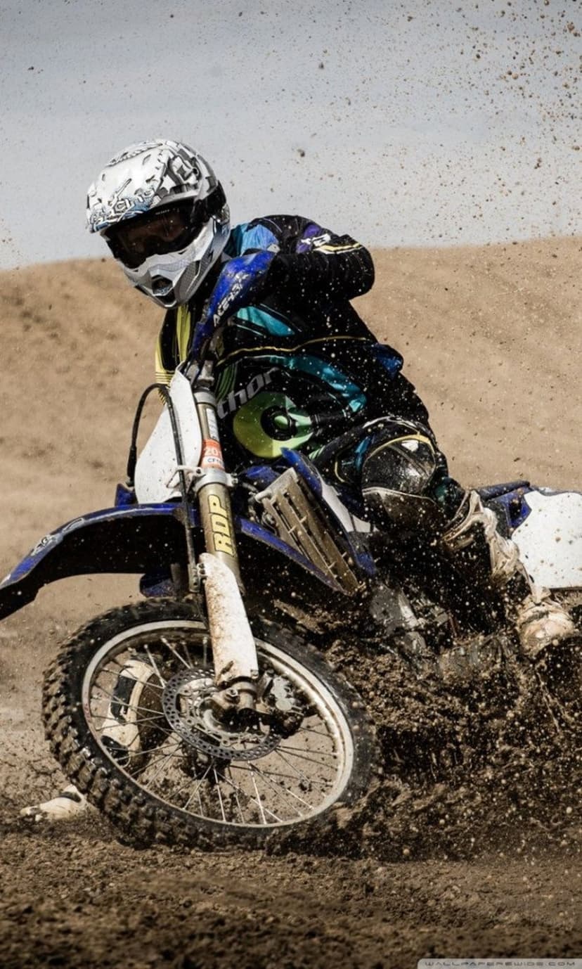 Motocross rider in full gear leans into a muddy track corner with dirt spraying from the rear motorcycle tire.