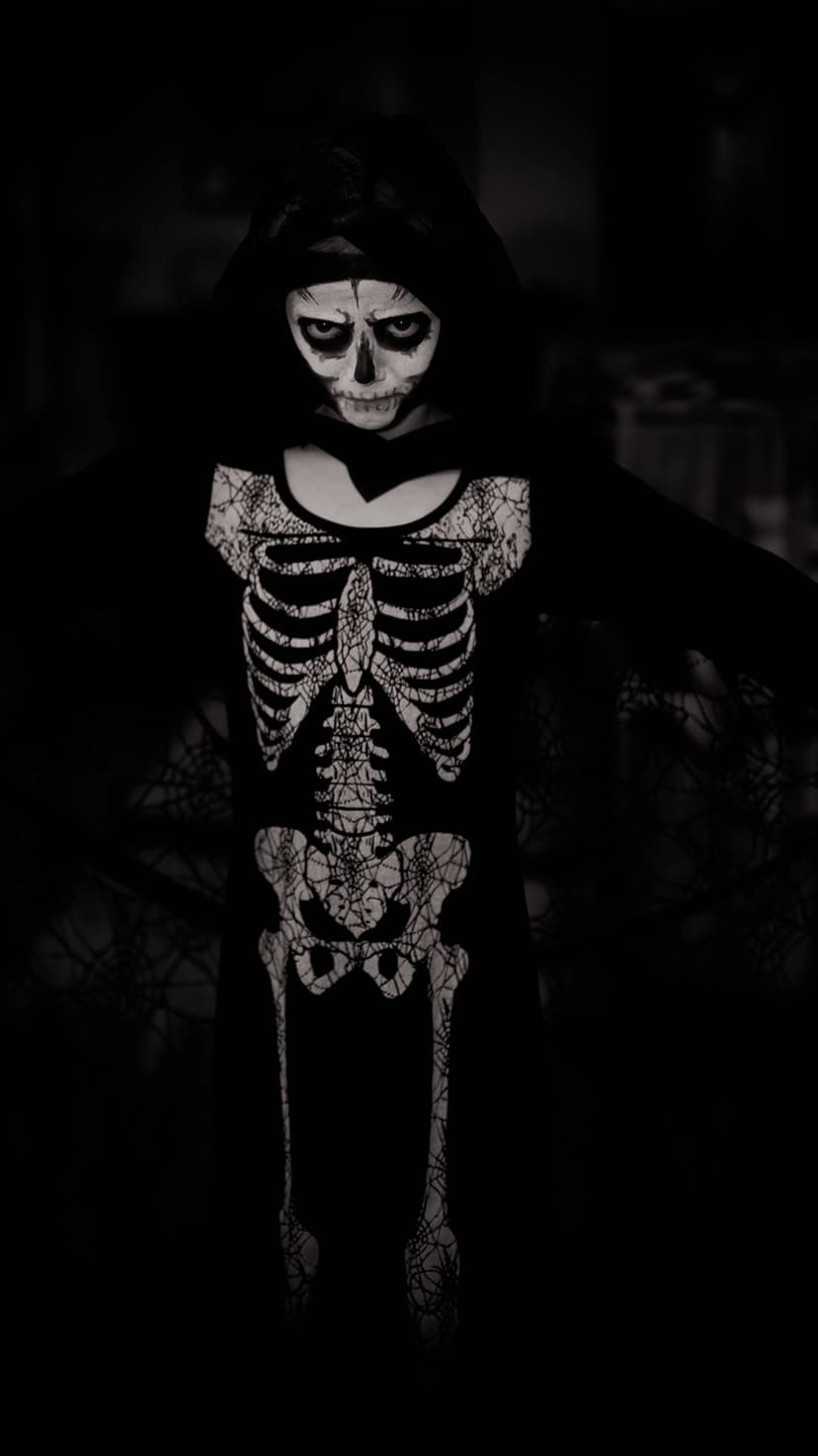 Child in a skeleton costume with detailed black and white face paint standing in dark, atmospheric lighting.